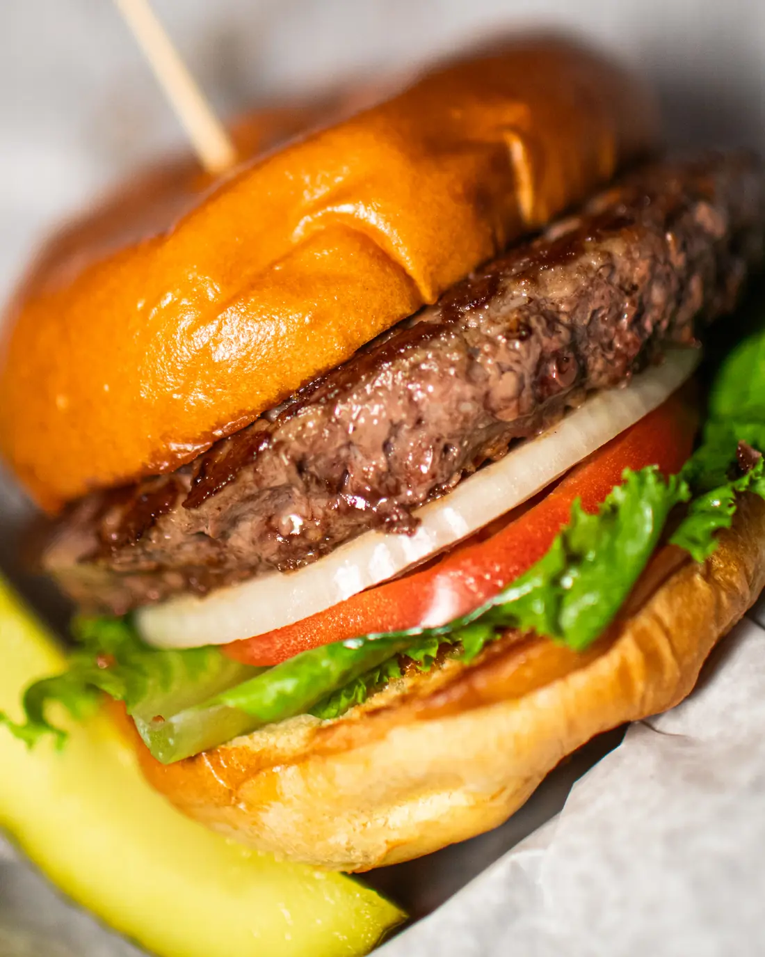 A burger on a brioche bun with lettuce, tomato and onion, pickle alongside.