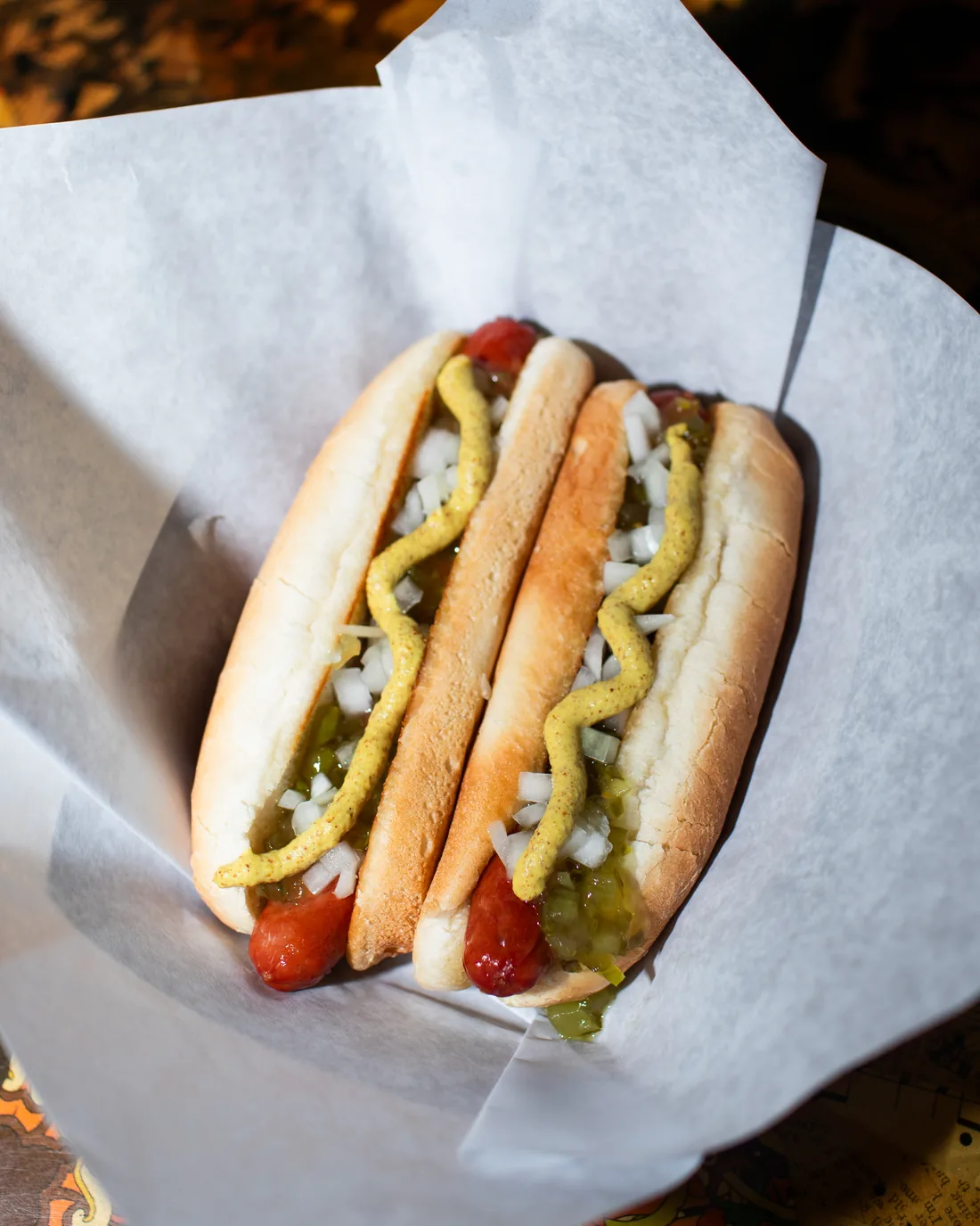 Two hot dogs in a paper-lined basket, split and topped with mustard, relish and chopped onion.