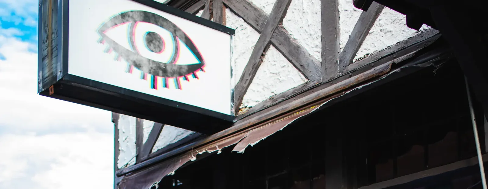 The Bond Street eye, hand-painted on a sign hung by bracket over the sidewalk, against the timbered facade and awning.